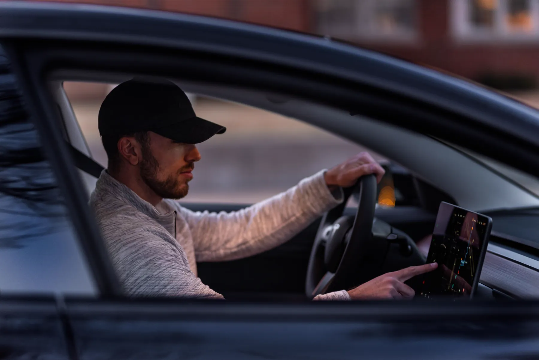 A man driving a Tesla, one hand on the wheel, seen through the side window